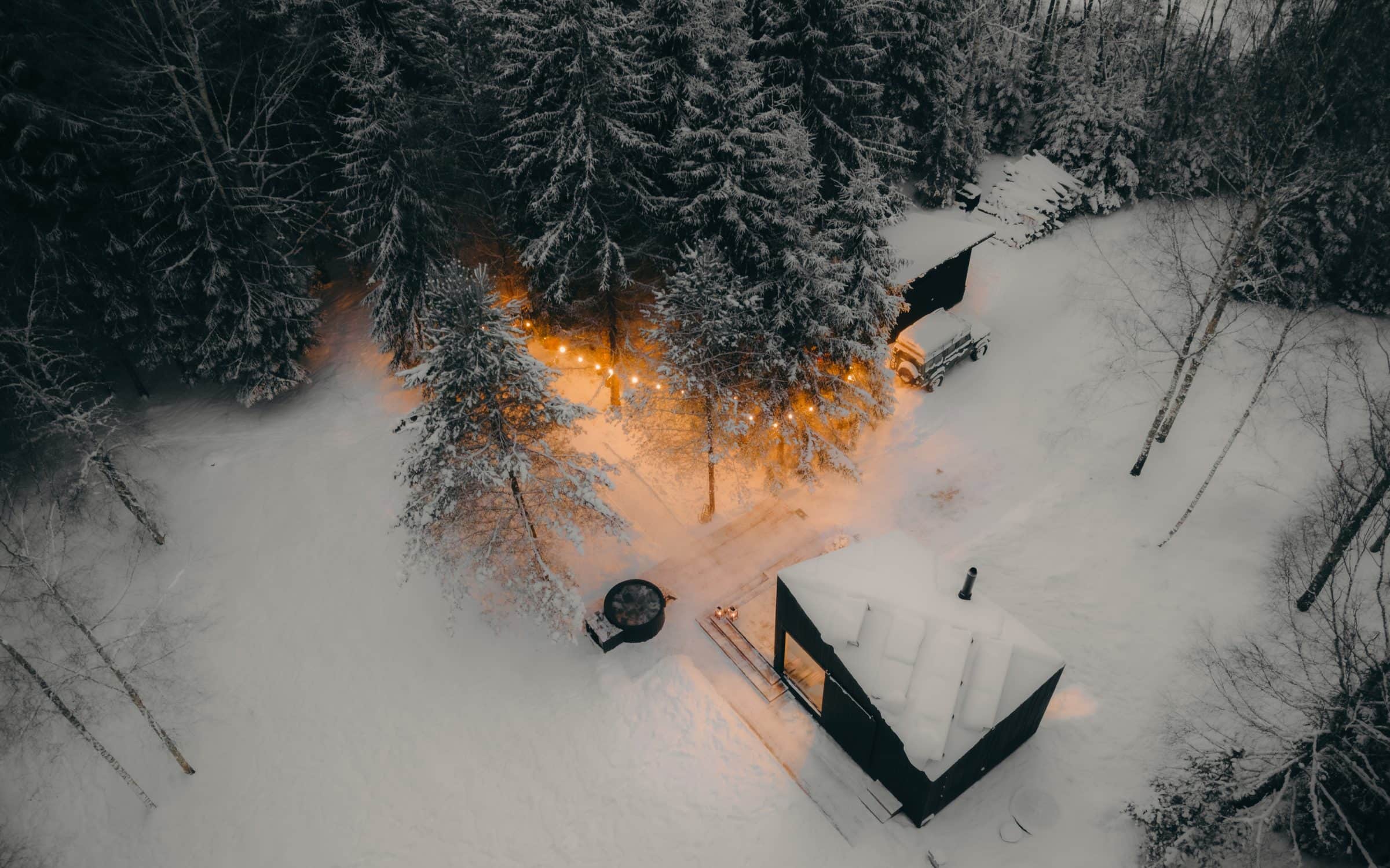 The cabin from above in winter — black roof, hot tub, string lights, snow
