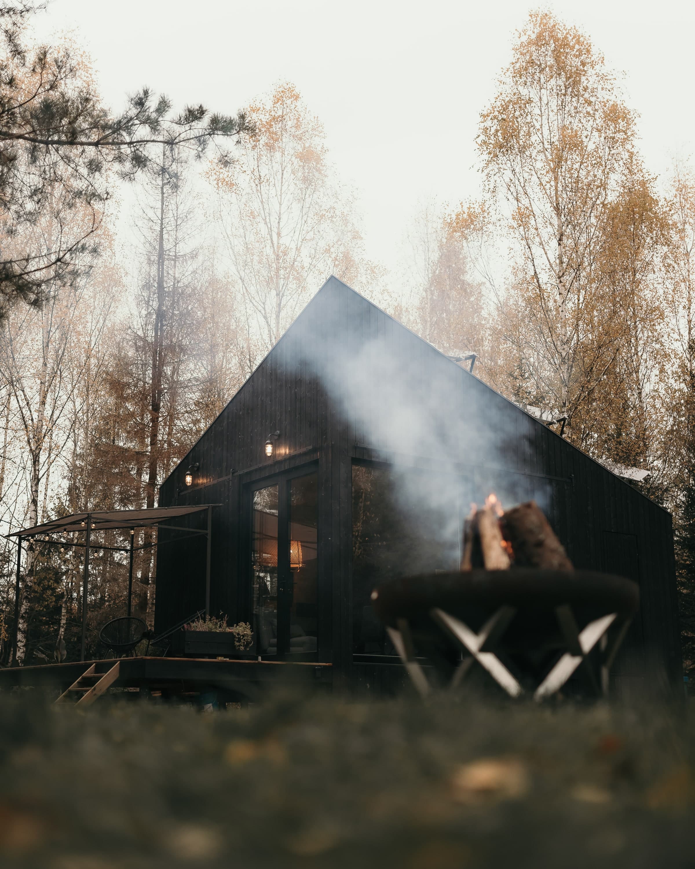 The cabin in autumn, smoke rising from the wood-stove chimney, fire pit in foreground
