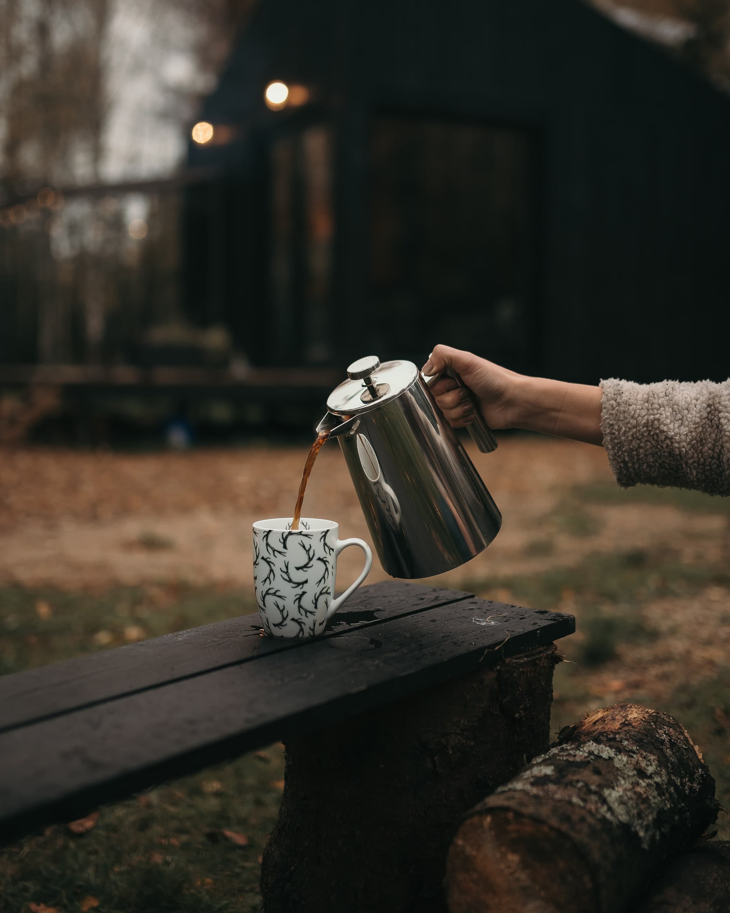 French press pouring coffee outside, cabin behind