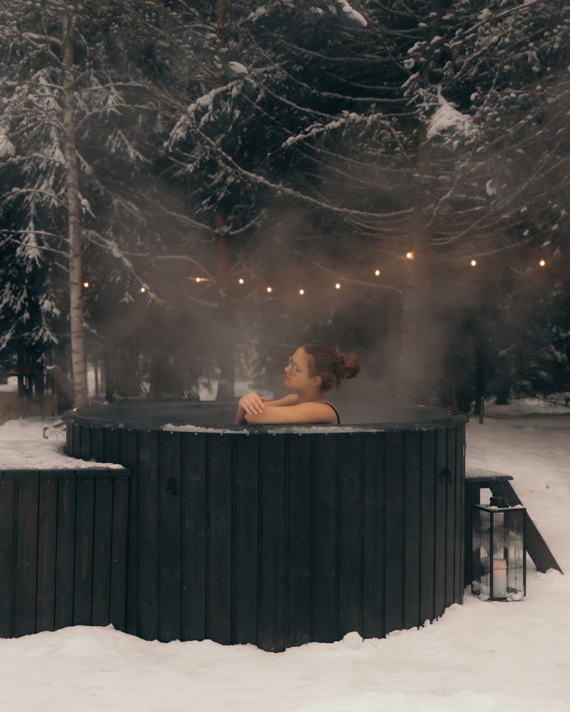 The hot tub in winter — snow firs, string lights, candle lantern